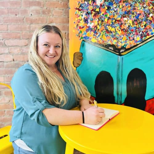 Woman writing at yellow table near colorful mural.